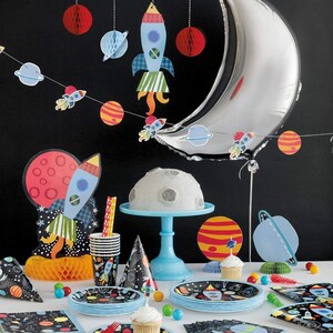May include: A space-themed party table setting with a silver crescent moon balloon, a cake shaped like a moon, and a rocket ship cake topper. The table is decorated with blue and black plates, cups, and napkins with a space theme. There are also red, orange, and blue paper decorations.