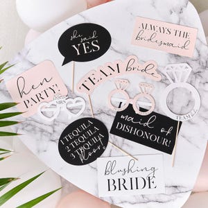 May include: A selection of photo booth props in pink, black, and white, perfect for a hen party. The props feature phrases such as "She said YES," "Hen Party," and "Always the Bridesmaid." Includes ring-shaped glasses and a diamond ring cutout.