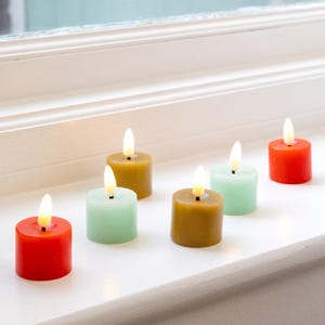 Op de afbeelding: Zes kleine, op batterijen werkende kaarsen in verschillende kleuren, waaronder rood, groen en goud. De cilindrische kaarsen hebben een flikkerend vlameffect, wat een warme sfeer creëert. Ze zijn op een witte vensterbank geplaatst. Hoogte: ongeveer 4 cm.