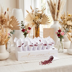 May include: A white box filled with paper cones containing red confetti, with the text "scatter some love." The image shows vases with dried flowers and roses, creating a celebratory atmosphere, ideal for a wedding or party.