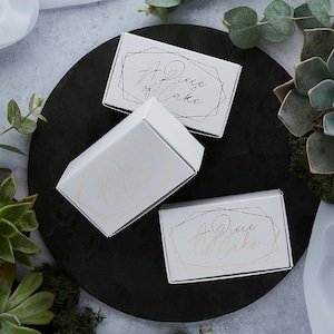 May include: Three white gift boxes with the text "A Piece of Cake" in a script font and geometric design. The boxes are arranged on a dark, round surface, surrounded by greenery. The text is in black and gold.