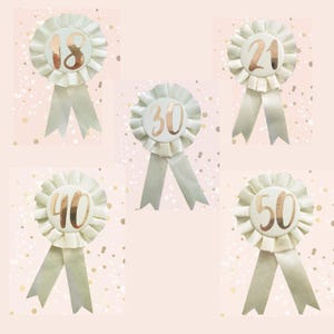 May include: Cream-colored rosette ribbons with gold numbers indicating ages 18, 21, 30, 40, and 50. Each ribbon has a pleated design and a matching cream-colored ribbon tail. The background is a light pink with gold confetti.