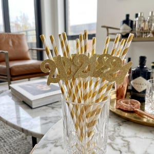 Può includere: Un bicchiere trasparente pieno di cannucce di carta a righe oro e bianche. Un topper glitterato dorato "2021" è attaccato alle cannucce. Le cannucce sono disposte su un tavolo di marmo, con una sedia in pelle e un bar sullo sfondo.