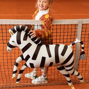 K&ouml;nnte beinhalten: Ein junges M&auml;dchen in einem braunen und gelben Cardigan mit Sonnenmuster h&auml;lt einen gro&szlig;en aufblasbaren Zebra-Ballon vor einem schwarzen Tennisnetz. Der Zebra-Ballon ist schwarz-wei&szlig; mit goldenen Akzenten.