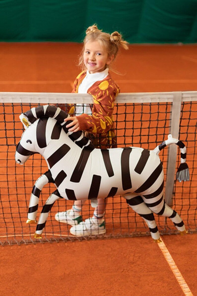 K&ouml;nnte beinhalten: Ein junges M&auml;dchen in einem braunen und gelben Cardigan mit Sonnenmuster h&auml;lt einen gro&szlig;en aufblasbaren Zebra-Ballon vor einem schwarzen Tennisnetz. Der Zebra-Ballon ist schwarz-wei&szlig; mit goldenen Akzenten.