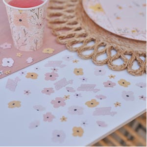 May include: A party scene with a floral theme. A paper cup with a pink drink sits next to a pink placemat and a woven placemat. Scattered across the white surface are flower-shaped confetti and text that reads "Happy Birthday".