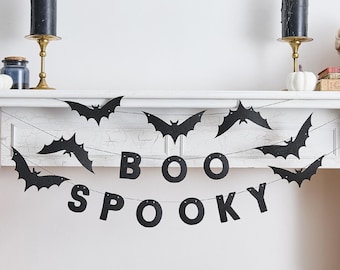 May include: A Halloween mantelpiece display with a black bat garland and a "BOO SPOOKY" banner. Two black candles in gold holders are on either side. A small jar and a white pumpkin are also visible.