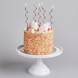 May include: A decorated cake with colourful sprinkles, macarons, and wavy pink candles. The cake is on a white cake stand against a grey backdrop. The candles are lit, with flames. The cake is round.