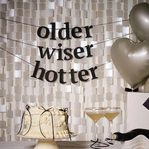 May include: A celebratory scene with a white cake, champagne glasses, and silver balloons. A banner reads "older wiser hotter" in black letters. The background is a wall of small, square, reflective tiles.