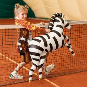 K&ouml;nnte beinhalten: Ein junges M&auml;dchen in einem braunen und orangefarbenen Pullover mit Sonnenmuster spielt auf einem Tennisplatz mit einem gro&szlig;en aufblasbaren Zebra-Spielzeug. Das Zebra ist schwarz-wei&szlig; mit goldenen Akzenten.