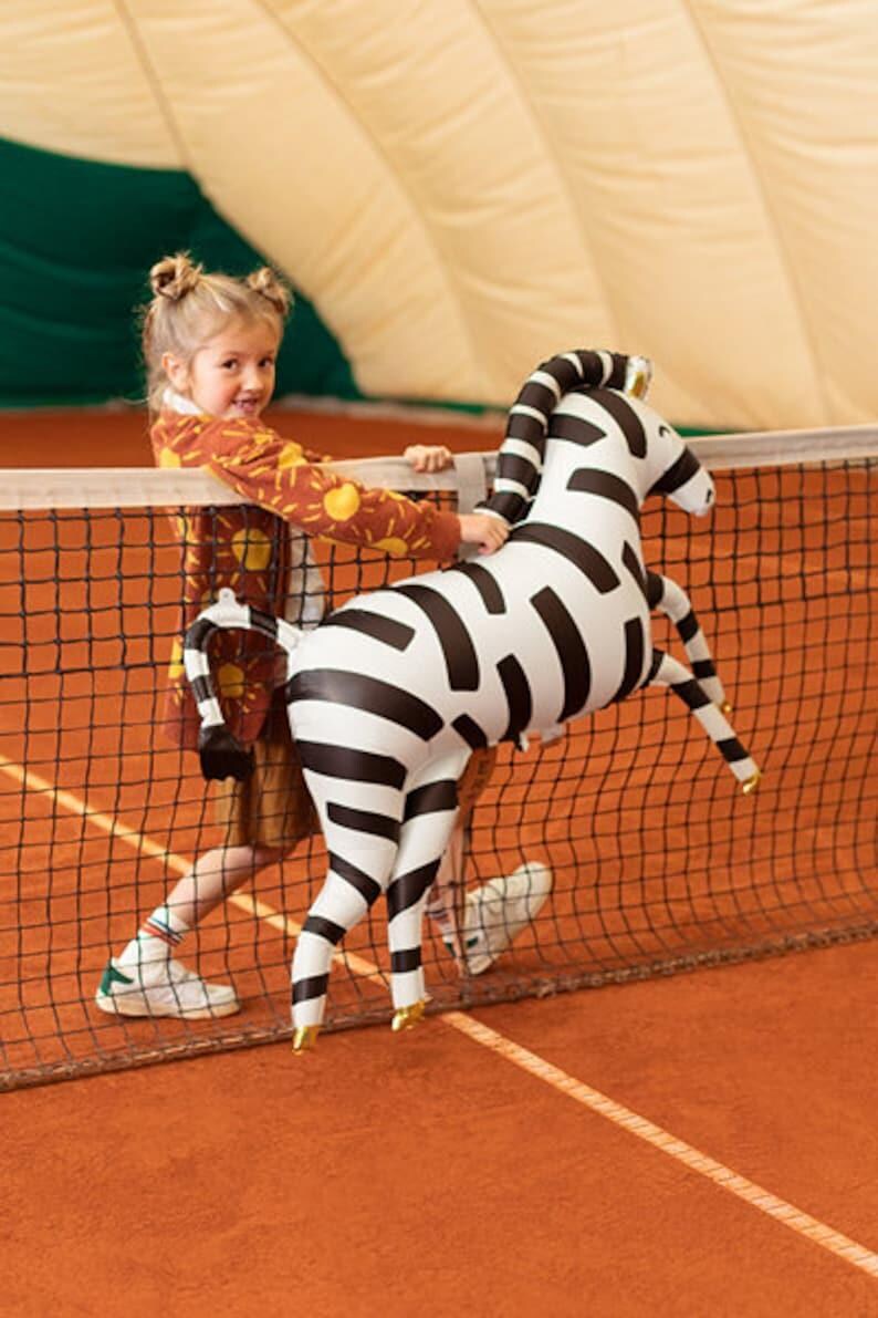 K&ouml;nnte beinhalten: Ein junges M&auml;dchen in einem braunen und orangefarbenen Pullover mit Sonnenmuster spielt auf einem Tennisplatz mit einem gro&szlig;en aufblasbaren Zebra-Spielzeug. Das Zebra ist schwarz-wei&szlig; mit goldenen Akzenten.