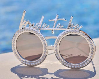 May include: Silver-toned sunglasses with round lenses and a decorative frame. The frame is embellished with small, round, pearl-like accents. The words "Bride to be" are written across the top of the frame in a cursive font.