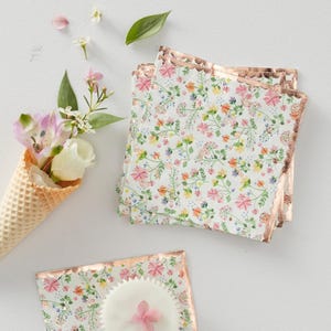 Op de afbeelding: Bloemenprint servetten met een roségouden rand, gestapeld naast een versierde cupcake en een wafelkegel gevuld met bloemen. De servetten hebben een delicaat patroon van roze, gele en blauwe bloemen op een witte achtergrond.