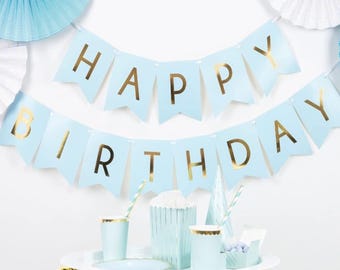 May include: A light blue "Happy Birthday" banner with gold lettering is displayed above a table of party supplies. The table includes cups, a popcorn container, and a small box, all in matching light blue and gold. Decorative paper fans are in the background.