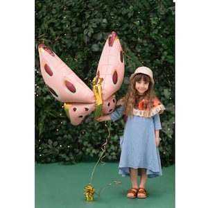 May include: A young girl wearing a blue dress and a straw hat holds a large pink and gold butterfly balloon. The balloon has brown spots and a gold glitter accent. The girl is standing in front of a green leafy background.
