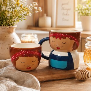 May include: Two ceramic mugs with a whimsical design. One is a small bowl, the other a larger mug with a blue handle. Both feature a brown-haired woman with a red polka-dot headband, closed eyes, and a red lip. The mugs are on a wooden surface.