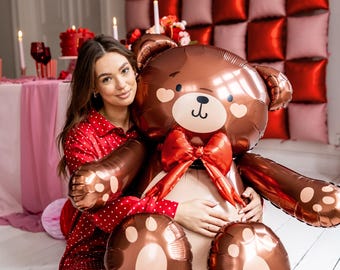 Può includere: Un grande palloncino a forma di orsetto marrone con un fiocco rosso, dettagli a forma di cuore e impronte di zampe. Il palloncino fa parte di una decorazione a tema San Valentino o romantico. Lo sfondo include palloncini rossi e rosa e decorazioni a cuore.