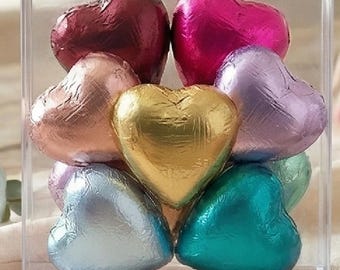 May include: A clear acrylic box filled with foil-wrapped heart-shaped chocolates. The chocolates are in various colors, including gold, pink, red, teal, silver, and lavender. The box is square and the chocolates are stacked inside.