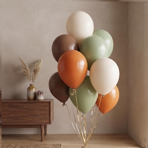 Może przedstawiać: Pęk okrągłych balonów w palecie barw kremowej, szałwiowej zieleni, ciemnego brązu i palonej pomarańczy. Balony są związane cienkimi, metalicznymi złotymi wstążkami, które zwisają w dół, tworząc dekoracyjną kompozycję unoszących się w powietrzu kul.