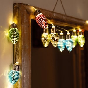 Può includere: Stringa di lampadine decorative con un design a mosaico. Le lampadine sono di vari colori, tra cui rosso, verde, giallo e blu. Le luci sono collegate da un filo sottile e sono appese vicino a una cornice di legno.