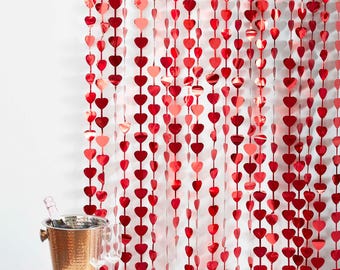 May include: Red heart-shaped garland hanging as a backdrop. The garland is made of multiple strands of red hearts, with a champagne bucket and glasses in the foreground. The hearts vary in shade from light to dark red.