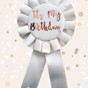 May include: A white birthday rosette with a circular center and two long ribbons. The center reads "It's My Birthday" in rose gold script. The rosette has a pleated design and is set against a light pink background with gold confetti.