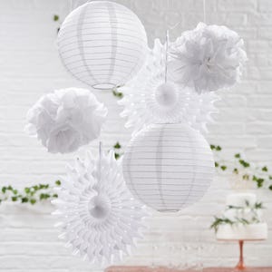 May include: A collection of white paper lanterns and decorative fans suspended from above. The lanterns are spherical with horizontal lines. Fluffy paper pom-poms and starburst-shaped fans provide texture. A white brick wall and cake are in the background.