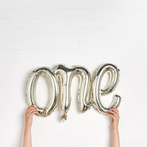 Może przedstawiać: Srebrny balon foliowy z napisem "one". Balon jest trzymany na tle białej ceglanej ściany. Balon jest dekoracją na przyjęcie z okazji pierwszych urodzin.