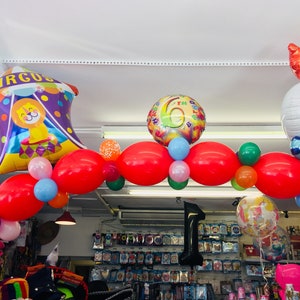 May include: A colorful balloon arch with red, green, blue, and pink balloons. The arch is decorated with a large purple and yellow circus tent balloon, a white balloon with a smiley face, and a silver balloon with the number 6 on it. The text "CIRCUS" is printed on the circus tent balloon.