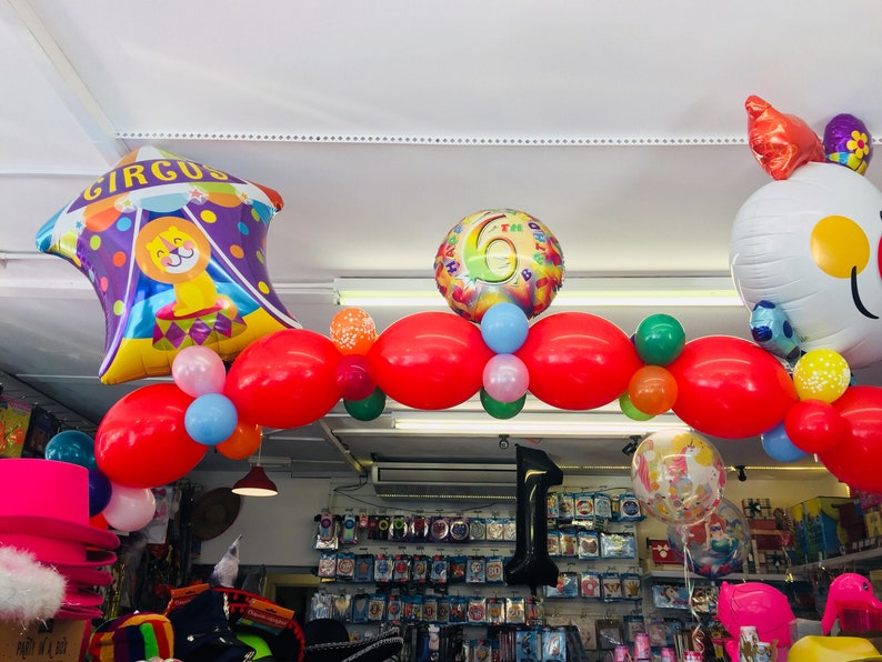 May include: A colorful balloon arch with red, green, blue, and pink balloons. The arch is decorated with a large purple and yellow circus tent balloon, a white balloon with a smiley face, and a silver balloon with the number 6 on it. The text "CIRCUS" is printed on the circus tent balloon.