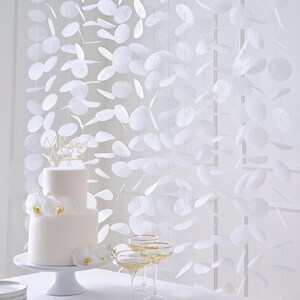 May include: A two-tiered white wedding cake with orchid decorations and a gold "Mr and Mrs" cake topper. Champagne glasses are in the foreground. A backdrop of hanging white paper shapes adds to the wedding theme.