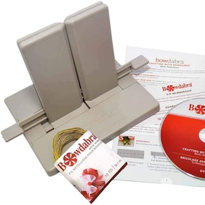 May include: A Bowdabra bow-making tool kit, including a gray plastic bow maker, a spool of gold bow wire, a DVD, and instructional papers. The DVD is labeled "Crafting with Bowdabra". The kit is designed for crafting bows and other decorative projects.