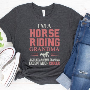 May include: A dark gray t-shirt with the text "I'm a Horse Riding Grandma" in red and white. The text is surrounded by a black line and a small horse silhouette. The text "Just like a normal grandma except much cooler" is printed below the main text.