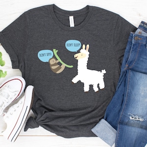 May include: A dark gray t-shirt with a white llama and a brown sloth on a green branch. The llama has a speech bubble that says "Don't Sleep!" and the sloth has a speech bubble that says "Don't Spit!"