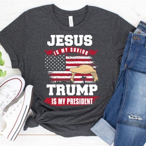 May include: A dark gray t-shirt with the text "JESUS IS MY SAVIOR TRUMP IS MY PRESIDENT" printed on it. The text is in white and red. An American flag is behind the text. The flag is distressed and has a white star on a blue background. A blonde hair wig is on top of the flag.
