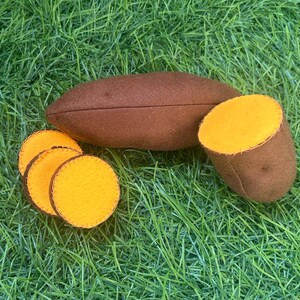 May include: A brown felt sweet potato with a sliced section and three round slices, showcasing a bright yellow interior. The felt food items are arranged on a bed of green artificial grass.