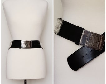 May include: A black leather belt with a silver buckle featuring an ornate design. The buckle is large and has a detailed image of an elephant.