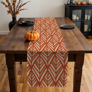 May include: A fall-themed table runner with a repeating leaf pattern in shades of orange, beige, and brown. The runner is placed on a dark wooden table, with a pumpkin and black plates adding to the autumn aesthetic.