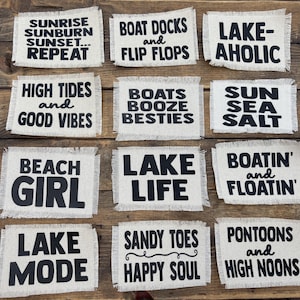 May include: A collection of twelve rectangular, off-white fabric coasters with black text. The coasters feature phrases like "Sunrise Sunburn Sunset... Repeat," "Lake Life," and "Sandy Toes Happy Soul," perfect for beach or lake house decor.