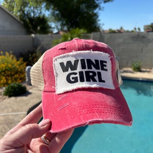 May include: A distressed pink baseball cap with a mesh back and a white rectangular patch on the front. The patch features the black text WINE GIRL in a bold, sans-serif font. The cap has a curved brim with visible frayed edges and stitching.