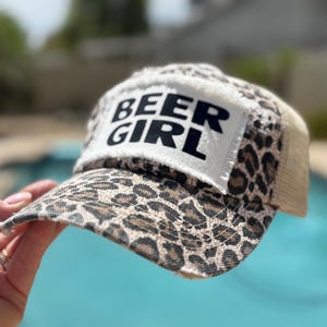 May include: A trucker hat with a leopard print pattern on the front and brim, and a white patch with the words "BEER GIRL" in black. The hat has a beige mesh back.