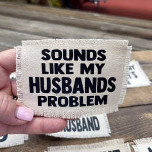 May include: A beige fabric patch with the phrase "SOUNDS LIKE MY HUSBANDS PROBLEM" printed in bold black letters. The patch has a frayed edge and is held up by a hand.