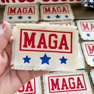 May include: A close-up of a hand holding a white fabric patch with the text "MAGA" in red letters and three blue stars. The patch has a frayed edge.
