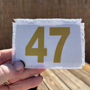 May include: A white rectangular fabric patch with frayed edges, displaying the number "47" in gold. The patch is held in a hand, with a blurred wooden background.
