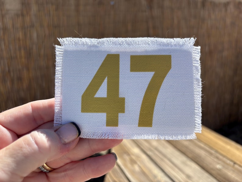 May include: A white rectangular fabric patch with frayed edges, displaying the number "47" in gold. The patch is held in a hand, with a blurred wooden background.