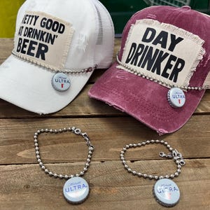 May include: Two trucker hats, one white and one burgundy, with the phrases "Pretty Good at Drinkin' Beer" and "Day Drinker" printed on them. Each hat has a bottle cap charm and a matching bracelet. The hats are on a wooden surface.