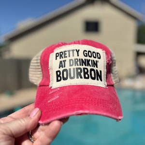 May include: A distressed red trucker hat with a white patch that reads "PRETTY GOOD AT DRINKIN' BOURBON" in black block letters. The hat has a mesh back and is held up against a blurred background.