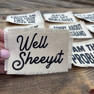 May include: Several small, rectangular fabric patches with black text on a tan background. One patch reads "Well Sheeyit" in a cursive font. Other patches have phrases like "Excuse Me, You Look Like You Love Me."