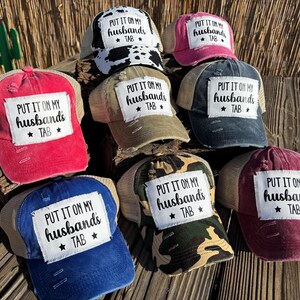 Peut inclure: Plusieurs casquettes de camionneur de couleurs variées, dont rouge, bleu et rose. Chaque casquette arbore un patch vieilli avec le texte "PUT IT ON MY husbands TAB" en lettres noires.