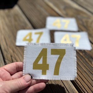 May include: A hand holds a rectangular patch with the number 47 printed in gold. The patch is white with a frayed edge. Three other patches with the number 47 are in the background.